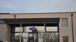 Der Eingang eines Gebäudes mit dem Namen AMS auf einer Glastür. Der Eingang hat ein modernes Design mit einem schwarzen Vordach und blauem Rand. Es gibt zwei Rauchverbotsschilder an den Glastüren.