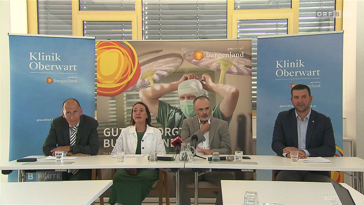 Eine Pressekonferenz mit vier Personen, die an einem Tisch sitzen, von denen einer ein Mann in grüner OP-Kleidung ist, der eine Maske aufsetzt.