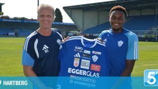 Zwei Männer in blauen Trikots, einer hält ein blaues Trikot mit mehreren Sponsorenlogos, stehen in einem Sportstadion mit leeren blauen Tribünen im Hintergrund.