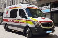 A white Mercedes-Benz ambulance with red and yellow stripes is parked on a city street. The ambulance has a red cross symbol on the side and a license plate on the front.