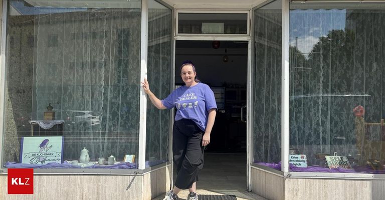 Eine Frau steht vor einem Geschäft mit einem lila T-Shirt, auf dem 'Cage Dealer' steht. Sie lächelt und lehnt sich gegen die Tür. Das Geschäft hat Glasfenster mit weißen Vorhängen und einem Schild, auf dem 'Keine Einnahmen gleich' steht. Es gibt Gegenstände zur Schau in der Fensterscheibe.
