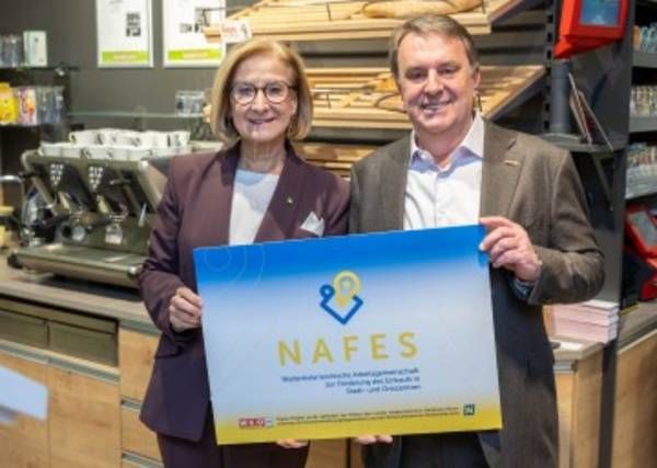 A man and a woman in business attire stand in a bakery, holding a large sign that reads 'NAFES'. Behind them, there is a coffee machine and shelves with bread.