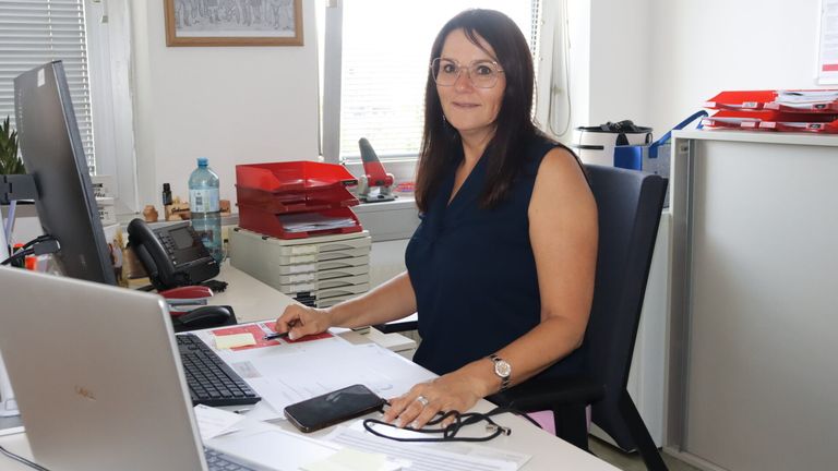 Eine Frau mit Brille und Uhr sitzt lächelnd in einem Büro an einem Schreibtisch, mit Tastatur, Telefon und Papieren vor sich.
