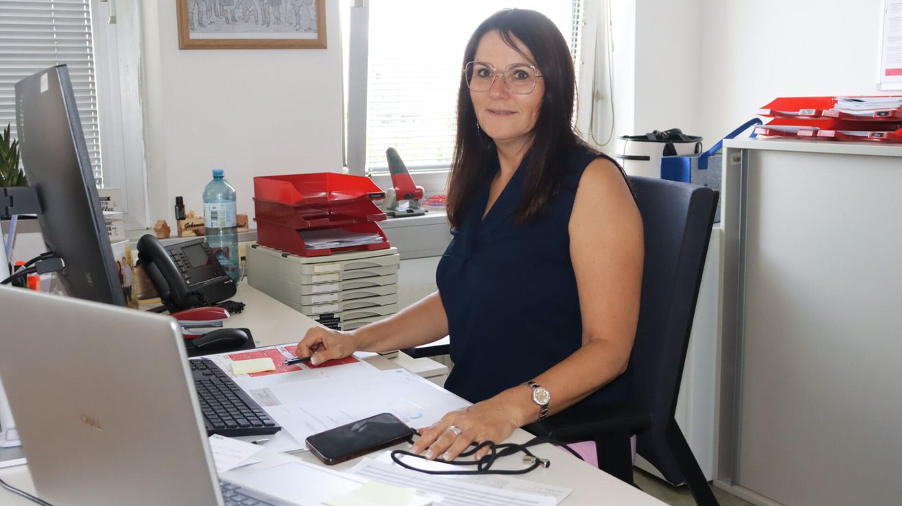 Eine Frau mit Brille und Uhr sitzt lächelnd in einem Büro an einem Schreibtisch, mit Tastatur, Telefon und Papieren vor sich.