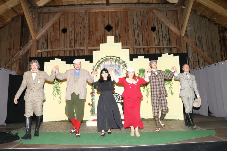 Eine Gruppe von Darstellern in Vintage-Kleidung hält auf einer Bühne mit gelbem Hintergrund und Holzdecke die Hände.