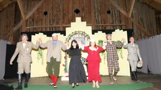 Eine Gruppe von Darstellern in Vintage-Kleidung hält auf einer Bühne mit gelbem Hintergrund und Holzdecke die Hände.