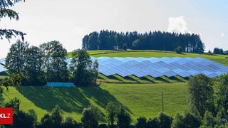 Eine große Solaranlage befindet sich auf einem grünen Hügel mit einem Wald im Hintergrund unter einem teilweise bewölkten Himmel.