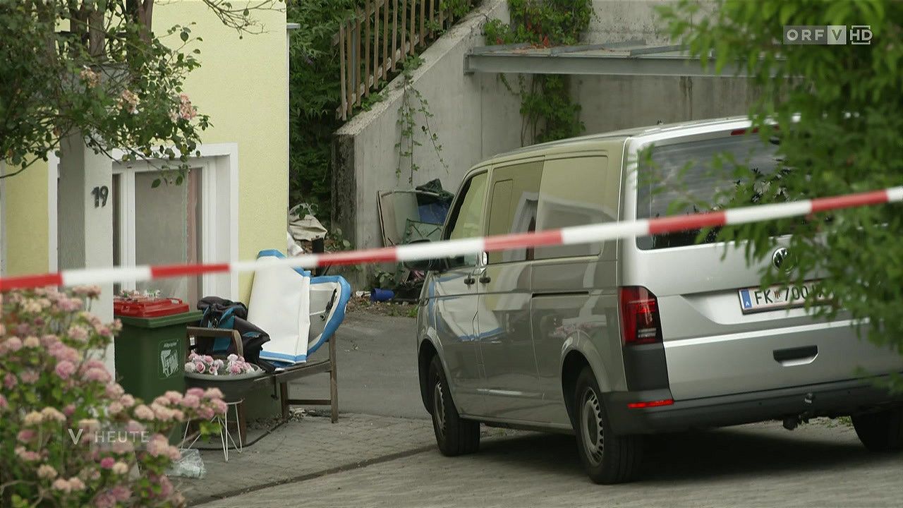 Ein silberner Lieferwagen ist am Straßenrand geparkt, teilweise durch ein rotes und weißes Absperrband blockiert. Ein Stuhl mit blauer und weißer Abdeckung steht in der Nähe.