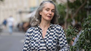Eine Frau mit grauem Haar trägt ein blau-weiß kariertes Hemd und eine Halskette auf der Straße.