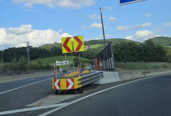 Eine Autobahn mit einem gelben und roten Schild, das einen Spurwechsel anzeigt, einem fahrenden Auto auf der Straße und einer Metallbarriere an der Seite.