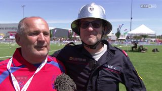 Zwei Männer stehen auf einem Feld mit einem unscharfen Hintergrund eines Sportereignisses. Der Mann auf der linken Seite trägt ein rotes Trikot mit einem Logo und einem Mikrofon. Der Mann auf der rechten Seite trägt eine Feuerwehruniform mit Helm.