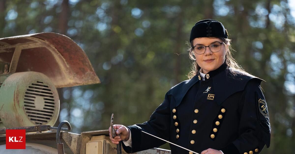 Eine Frau in einer dunkelblauen Uniform mit goldenen Knöpfen und einer schwarzen Mütze hält einen Dirigentenstab und lächelt in die Kamera. Sie steht vor einer Maschine mit einem rostigen Metallteil.