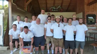 Eine Gruppe von Männern in weißen T-Shirts und verschiedenen Shorts posiert für ein Foto in einem Raum mit Holzbalken und einem Fahrrad an der Decke.