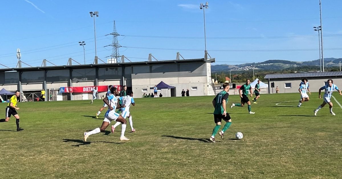 Fußballspieler in grünen und weißen Trikots rennen auf einem Feld. Der Spieler in Grün tritt den Ball. Das Feld ist von einem Gebäude mit einem roten Banner umgeben.