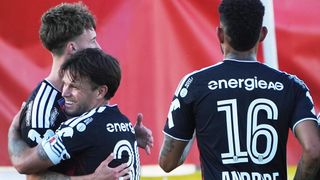 Drei Fußballspieler feiern ein Tor, einer trägt ein Trikot mit der Nummer 3 und ein anderer die Nummer 16. Der Spieler in der Mitte lächelt und umarmt den Spieler auf seiner linken Seite.