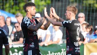 Zwei Fußballspieler feiern ein Tor mit einem High-Five vor einem Publikum. Einer trägt ein schwarzes Trikot mit 'Kiso' und der andere hat 'energie' auf seinem.