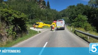 Ein gelber Hubschrauber und ein Krankenwagen sind am Straßenrand geparkt, wobei Personen in roten Uniformen einen Patienten versorgen.