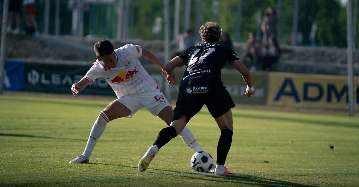 Zwei Fußballspieler sind auf einem Feld in Aktion, einer trägt ein weißes Trikot mit einem roten Bullenlogo und der andere in Schwarz mit der Nummer 12. Sie sind beide in Bewegung, einer versucht den Ball zu treten, während der andere verteidigt.