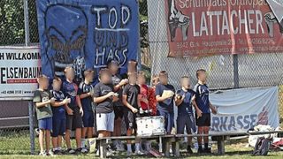 Eine Gruppe junger Jungen, möglicherweise eine Marschkapelle, steht vor einem Zaun mit Bannern. Sie tragen Uniformen und einige spielen eine Trommel.