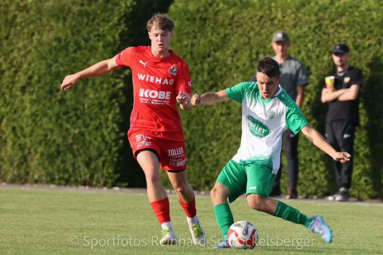 Zwei Fußballspieler in roten und grünen Trikots sind in einem Spiel, einer versucht, dem anderen den Ball zu stehlen. Dahinter beobachtet ein Mann in einer Mütze.