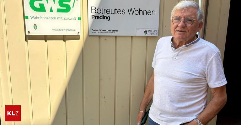 Ein älterer Mann steht vor einem Gebäude mit einem Schild, das 'Betreutes Wohnen Preding' besagt. Er trägt ein weißes Hemd und hält ein grünes Objekt, möglicherweise ein Werkzeug.