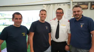 Vier Männer stehen zusammen auf einem Gruppenfoto, zwei tragen dunkelblaue T-Shirts mit Logos, einer ein weißes Uniformhemd mit schwarzer Krawatte und der letzte in einem blauen Hemd. Hinter ihnen ist ein weißes Zelt mit einem Schild zu sehen, das 'Wiener Schnitzel mit Salat 13' anzeigt.
