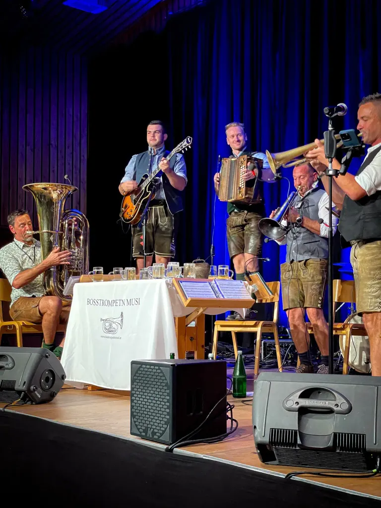 Fünf Männer spielen auf der Bühne mit Musikinstrumenten. Ein Mann spielt Gitarre, ein anderer Trompete und die anderen Tuba, Akkordeon und Schlagzeug.