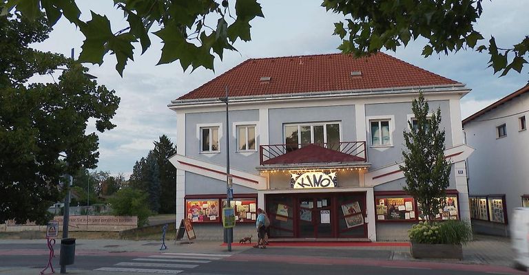 Ein Gebäude mit einem roten Dach und einem Schild, das 'KINO' steht. Zwei Personen gehen davor. Es gibt Pflanzen und Bäume um das Gebäude.