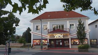 Ein Gebäude mit einem roten Dach und einem Schild, das 'KINO' steht. Zwei Personen gehen davor. Es gibt Pflanzen und Bäume um das Gebäude.