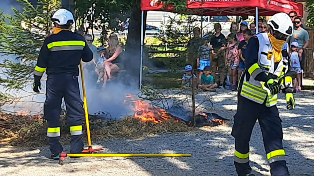 Ein Feuerwehrmann löscht ein Feuer mit einem Schlauch, während eine Gruppe von Menschen, darunter Kinder, aus der Ferne zusieht.