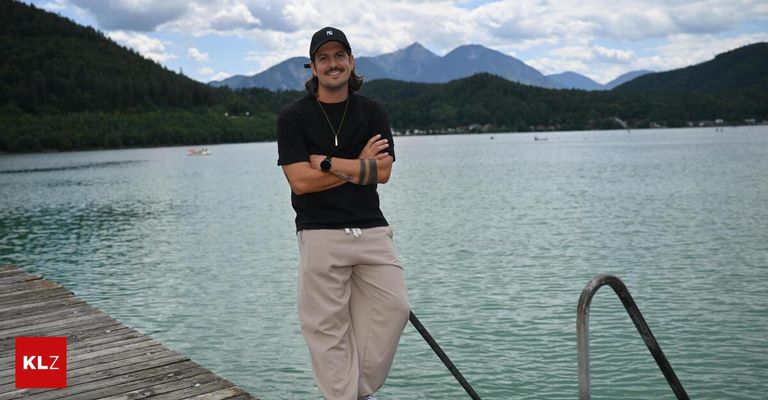 Ein Mann mit schwarzem Hut und Halskette steht auf einem Steg am See mit Bergen im Hintergrund.