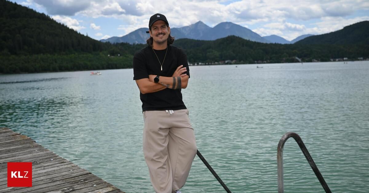 Ein Mann mit schwarzem Hut und Halskette steht auf einem Steg am See mit Bergen im Hintergrund.
