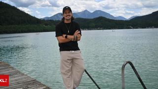 Ein Mann mit schwarzem Hut und Halskette steht auf einem Steg am See mit Bergen im Hintergrund.