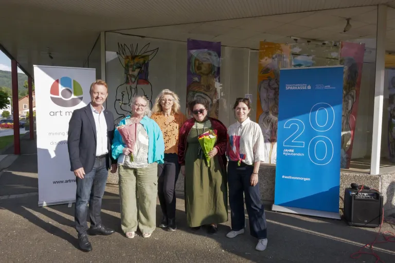 Fünf Personen stehen vor einem Gebäude mit einem blauen Banner, das '20 Jahre #glaubandiich' besagt. Die Frau in der Mitte hält einen Blumenstrauß. Hinter ihnen befinden sich Glaswände mit abstrakter Kunst.