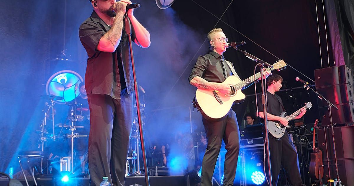 Zwei Männer treten auf der Bühne auf, einer singt in ein Mikrofon und der andere spielt Gitarre. Die Bühne ist mit blauen und weißen Lichtern beleuchtet.