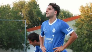Ein Fußballspieler in einem blauen Trikot mit weißen Besätzen steht mit den Händen auf den Hüften und schaut nach oben. Ein weiterer Spieler in einem ähnlichen Trikot steht hinter ihm. Sie befinden sich auf einem Spielfeld mit Bäumen und einem Gebäude im Hintergrund.