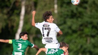 Ein Fußballspieler in einem weißen Trikot mit der Nummer 17 springt, um den Ball zu köpfen, während zwei andere Spieler in grünen Trikots versuchen, ihn zu blockieren.