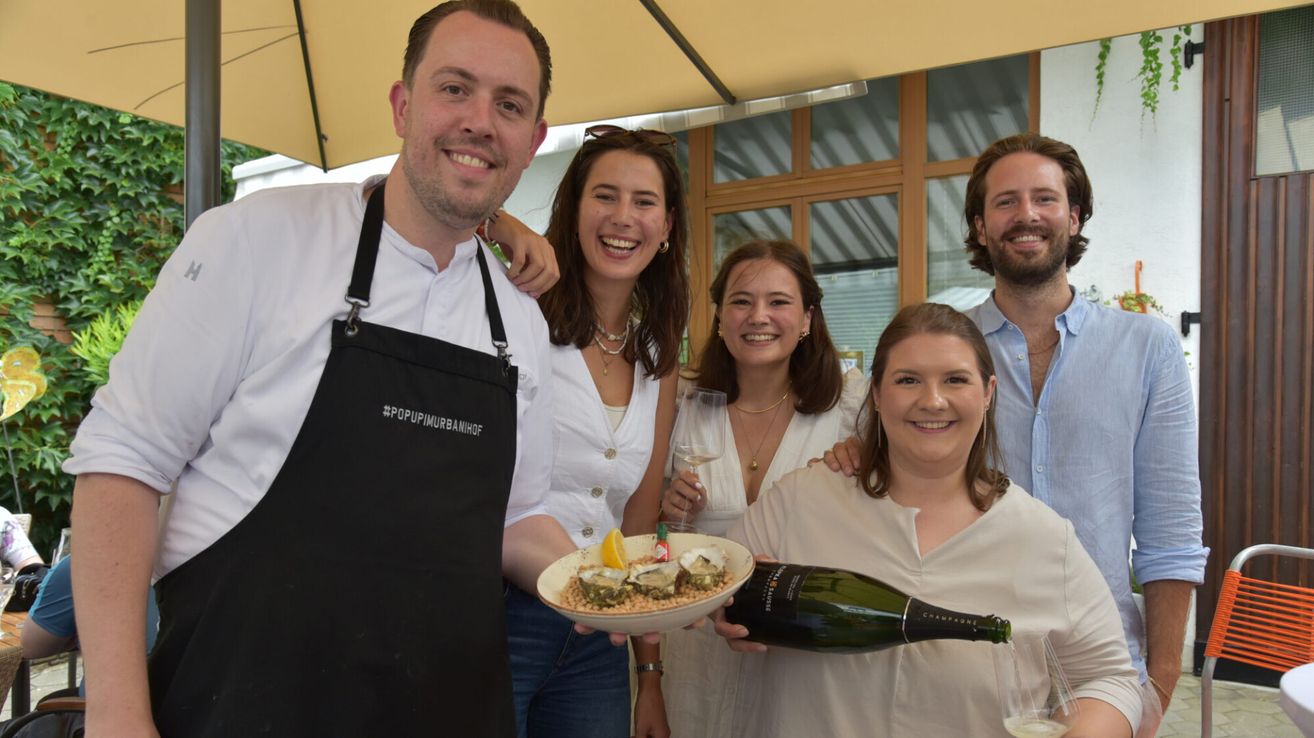 Eine Gruppe von Menschen lächelt und posiert für ein Foto. Ein Mann in einer schwarzen Schürze hält einen Teller mit Austern und eine Flasche Champagner. Eine Frau auf der rechten Seite hält eine Flasche Champagner. Hinter ihnen ist ein Glasfenster.