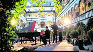 Ein klassisches Musikkonzert in einem prunkvollen Innenhof mit einem Flügel, einem Geiger und Publikum.