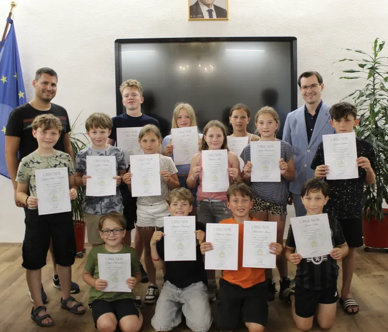 Eine Gruppe von Kindern und Erwachsenen posiert für ein Foto, während sie Urkunden halten. Sie stehen und knien auf einem Holzboden. Hinter ihnen befindet sich ein großer Monitor und ein Bilderrahmen an der Wand.
