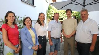 Vier Personen stehen zusammen in einer Außenumgebung und lächeln für ein Foto. Eine Frau in einer blauen Jacke und einem gelben Oberteil ist links, und eine Frau in einer weißen Bluse ist neben ihr. Zwei Männer sind rechts, einer in einem hellblauen Hemd und der andere in einem gestreiften Hemd.