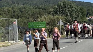 Eine Gruppe von Kindern in traditioneller bayerischer Kleidung, die an einem Umzug teilnehmen, mit einem grünen Schild im Vordergrund und einer Blaskapelle, die hinter ihnen marschiert.