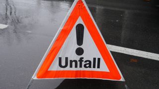 Ein dreieckiges Verkehrsschild mit orangefarbenem Rand und weißem Hintergrund. Es zeigt einen schwarzen Ausrufepunkt und das Wort 'Unfall' in fetten schwarzen Buchstaben, was auf einen kürzlichen Unfall hinweist. Das Schild steht auf einer nassen Straßenoberfläche.