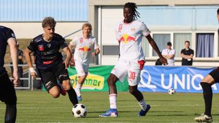 Drei Fußballspieler auf einem Feld, einer in einem weißen Trikot mit einem roten Bullenlogo, ein anderer in einem blauen Trikot und ein dritter in einem weißen Trikot mit einem roten Bullenlogo. Der Spieler in Weiß läuft auf den Ball zu.