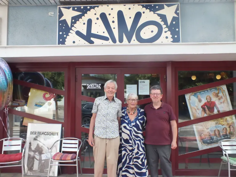 Drei Personen stehen vor einem Gebäude mit einem Schild, das 'KINO' steht. Der Mann links trägt ein Blumenhemd und Khakihosen, die Frau in der Mitte trägt ein blau-weißes Kleid und der Mann rechts ein Weinrotes Poloshirt. Hinter ihnen spiegelt eine Glastür ein Auto und ein Poster.