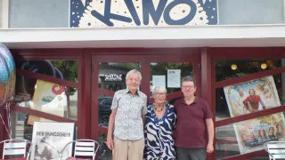 Drei Personen stehen vor einem Gebäude mit einem Schild, das 'KINO' steht. Der Mann links trägt ein Blumenhemd und Khakihosen, die Frau in der Mitte trägt ein blau-weißes Kleid und der Mann rechts ein Weinrotes Poloshirt. Hinter ihnen spiegelt eine Glastür ein Auto und ein Poster.