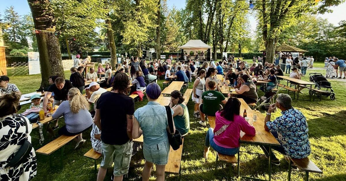 Eine große Menschenmenge versammelt sich in einem Park für eine Outdoor-Veranstaltung. Viele sitzen an Picknicktischen, während andere stehen und eine Bühnenaufführung beobachten. Die Umgebung ist von Bäumen und Grünflächen umgeben.