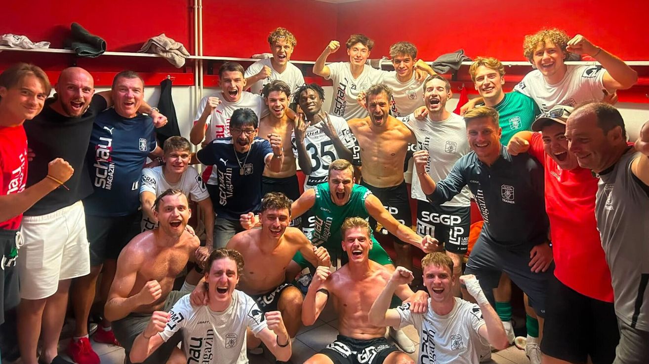 Eine Gruppe junger Männer in Sportkleidung, teilweise ohne Hemd, feiert in einer Umkleidekabine mit einer roten Wand im Hintergrund. Sie posieren für ein Foto mit verschiedenen Ausdrücken von Freude und Begeisterung.