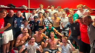 Eine Gruppe junger Männer in Sportkleidung, teilweise ohne Hemd, feiert in einer Umkleidekabine mit einer roten Wand im Hintergrund. Sie posieren für ein Foto mit verschiedenen Ausdrücken von Freude und Begeisterung.