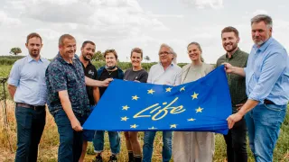 Eine Gruppe von Menschen, die eine blaue Flagge mit dem Wort 'Leben' und gelben Sternen in der Mitte halten, steht auf einem Grasfeld unter einem bewölkten Himmel.